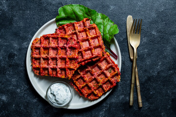 Vegetarian beetroot and zucchini waffles with herbs on a plate with sour cream on table, closeup, top view. Tasty vegetable waffles for eat made with waffle maker