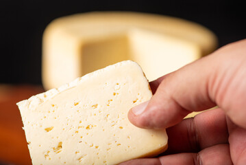 Semi-cured cheese made in Minas Gerais, Brazil, with a human hand holding a slice, on a rustic surface and dark background, selective focus.