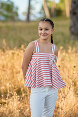 Smiling girl in a striped top poses in a sunlit field during golden hour with trees in the background