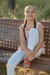 Smiling girl with braids sitting on a bench in a sunny park during late afternoon