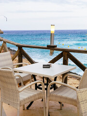A reserved restaurant table with wicker chairs overlooks the turquoise sea in Mallorca, with a wooden railing and soft lamp enhancing the peaceful coastal atmosphere