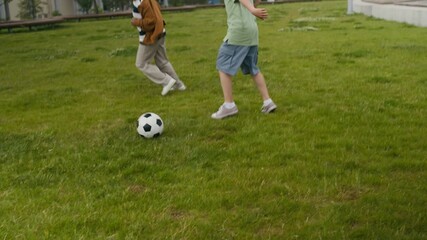 Two intercultural kids in casualwear running after soccer ball along green lawn or football field outdoors and kicking it while playing leisure sports game - Powered by Adobe