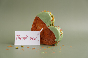 THANK YOU text paper note international Thanksgiving day gift Heart shaped pistachio croissant on green background with copy space. Puff pastry new trendy sweet dessert bakery. Creamy dough experiment