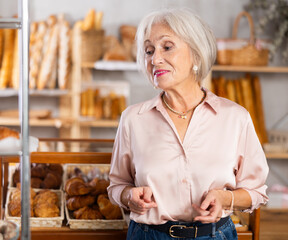 Senior woman looks at glass showcase filled with various flour products, bread assortment, croissants, puffs with filling. Quick snack on go, bakery, waiting for order
