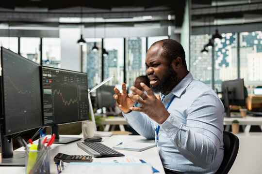 Trader in office raging after losing money, reacting negatively to poor portfolio performance. African american man upset by financial losses due to volatile market swings, yelling