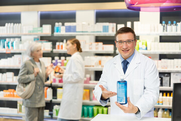 Polite middle-aged male pharmacist demonstrating lubricant in chemist's shop with large assortment...