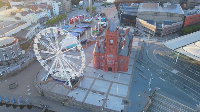 Cardiff Bay aerial shot over the Pierhead Building and ferris wheel and fair that reverses and ascends to show the city centre skyline in the background, people walk below, Cardiff, UK, 4K, 60fps
