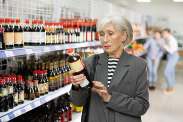 Interested focused senior female shopper choosing traditional Chinese soy sauce in Asian store, carefully reading labels on bottle..