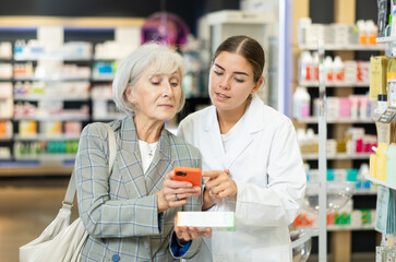 Concerned old woman showing to young female pharmacist QR-code of product on her smartphone in chemist's shop