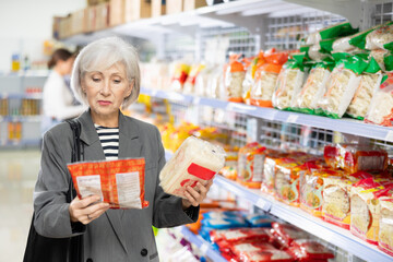 Senior woman in supermarket buys daily groceries, pasta Chinese noodles. Female pensioner buyer examines packaging of product and reads information on label