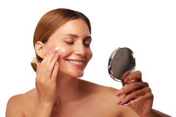 A woman joyfully applies cream to her face, gazing at her reflection in a handheld mirror. She...