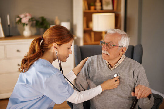 In a living room, a female home caregiver listens to an older gentleman's heartbeat with a stethoscope. The senior man is sitting in an armchair and holding a cane. - Powered by Adobe
