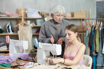 Annoyed elderly woman teaching young woman how to sew on machine in sewing workshop