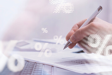Close-up of a hand holding a pen over financial documents with a laptop and calculator in the background, overlaid with percentage signs. Concept interest rate calculations and economic analysis.