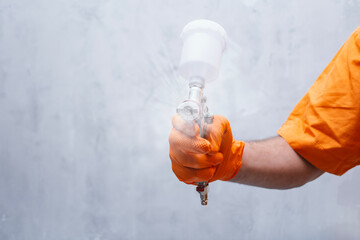 Close-up of painter hand in orange protective glove holding spray gun and applying paint. Car painting, industrial coating, body repair and maintenance concept.
