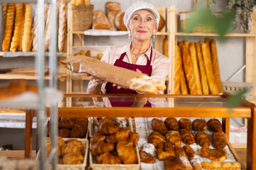 Positive mature woman bakery employee offering bread and different baguettes for sale