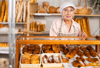 Selling fresh croissants in a bakery - smiling female baker places hot croissants on display