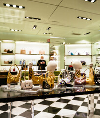 Blurred view of a luxury handbag store interior with shiny designer bags on display, checkered floor tiles, soft lighting and a masked employee