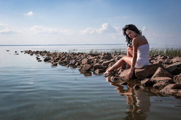 Young sexy beautiful woman is sitting on the rocks in the sea near the shore and enjoys relaxation, the warm sun and enjoys the sunset and touches the water with his hands. Wellness, tourism, vacation