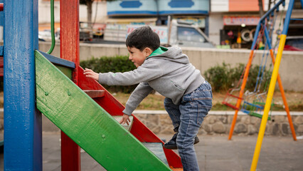 Little boy in gray hoodie and jeans climbing up a colorful wooden structure at a playground on a cloudy day.