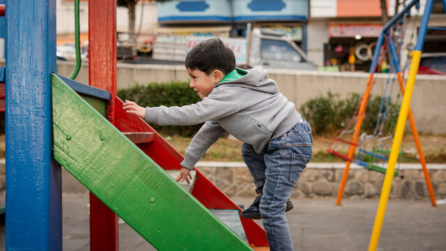 Little boy in gray hoodie and jeans climbing up a colorful wooden structure at a playground on a cloudy day.