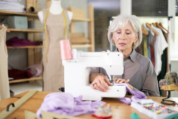 Older dressmaker works at a sewing machine against the background of racks with fabrics....