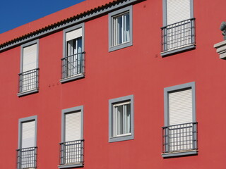  Facade of a building in a bright red color. The numerous windows are striking, some of which have closed shutters and others have small balconies with metal railings.