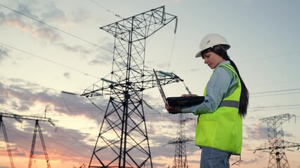 Woman engineer standing by power tower with laptop, electrical maintenance workflow, infrastructure monitoring outdoors, electricity grid managed by female expert, using digital tools at utility site