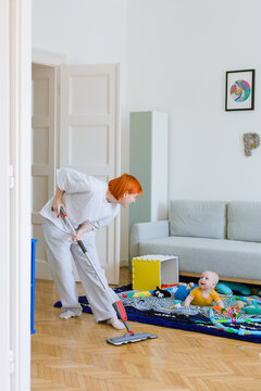 A woman mops the floor in a living room, a child is lying on carpet