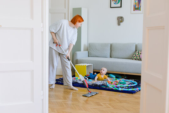 A woman mops the floor in a living room, a child is lying on carpet