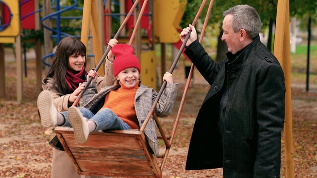 Baby daughter dad mom play together on swing in autumn park, family fun. Little girl parents swinging on swing. Dad mom child play on playground in city park. Happy family. Parents boy family weekend