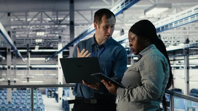 Team of engineers monitor server hub activity using laptop and tablet. Teamworking data center colleagues review rigs tests results on devices to ensure systems are working correctly, camera B