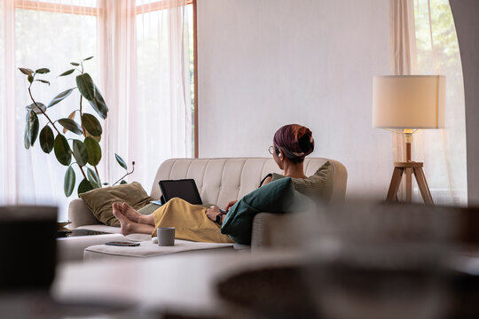 Woman relaxes on sofa with digital tablet in bright room.