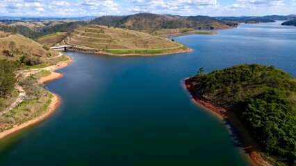 Impressionante paisagem aérea de barragem rodeada por colinas verdes e céu azul, perfeita para projetos turísticos e naturais.