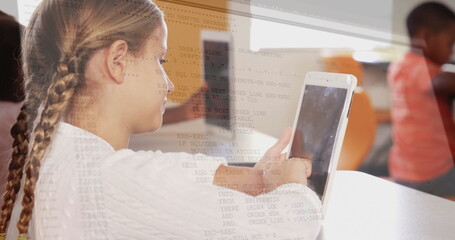 Schoolgirl sitting at school desk viewing code overlay on tablet computer, with blurred classmate