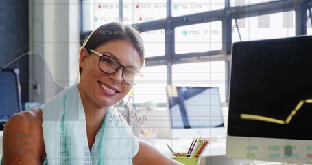 Reviewing financial overlay on screens mid-adult woman at desk in office, with pencil cup and pens