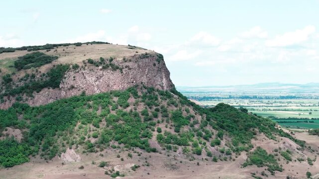 A majestic aerial drone shot orbits the Măgura Uroiului mountain in Romania. The view showcases the sheer rock cliff, green slopes, and the vast Transylvanian plain below.