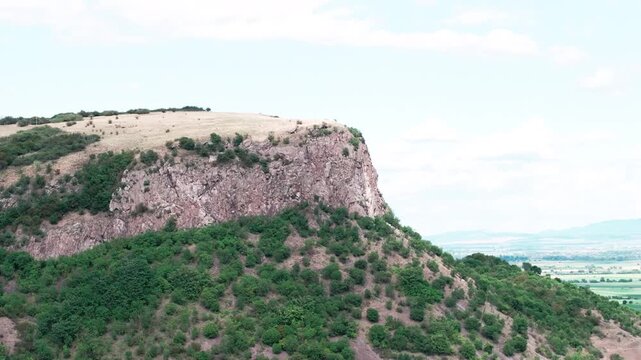 Aerial tele drone shot orbits the Măgura Uroiului mountain in Romania. The view showcases the sheer rock cliff, green slopes, and the vast Transylvanian plain below.