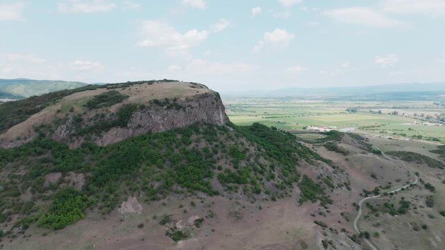 A super wide aerial drone shot makes a sweeping orbit around the majestic Măgura Uroiului mountain in Romania, revealing the sheer rock cliff and the vast surrounding plain.