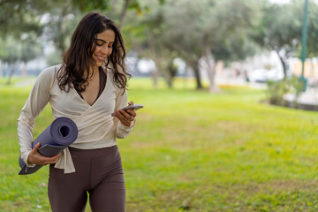 Yoga instructor using smartphone and carrying yoga mat in park