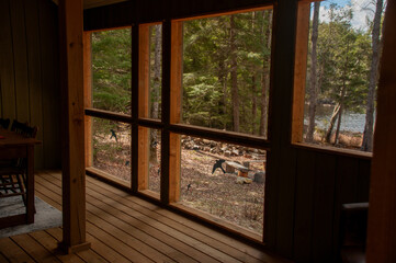 A cozy, rustic cottage interior with wooden floors and paneling, looking out through large windows to a wooded landscape and a sunlit lake.