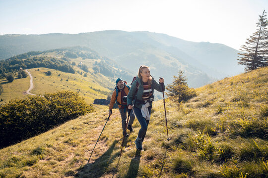 Couple hiking up a mountain hill with trekking poles on sunny day - Powered by Adobe