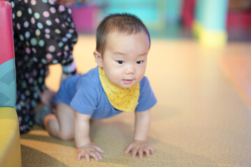 Fototapeta premium In an indoor play area, parents in their thirties watch a one-year-old baby crawl on a soft mat with warm smiles, toys nearby and gentle light in the room. Hongkou District, Shanghai, China. Spring.