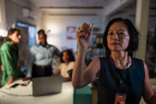 Teamwork, Woman writes math on glass board, others collaborate.