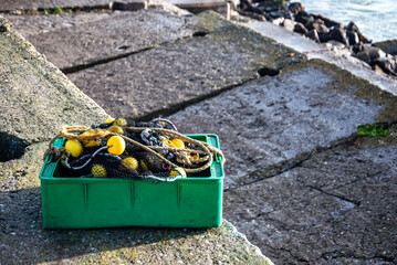 Green plastic box filled with fishing nets and yellow buoys placed on a concrete pier by the sea. Representative image of artisanal and commercial fishing activity.