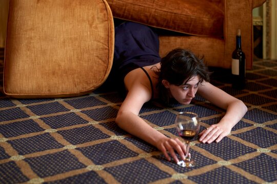 Young Woman Crawls on Floor Under Couch With Drink in Hand