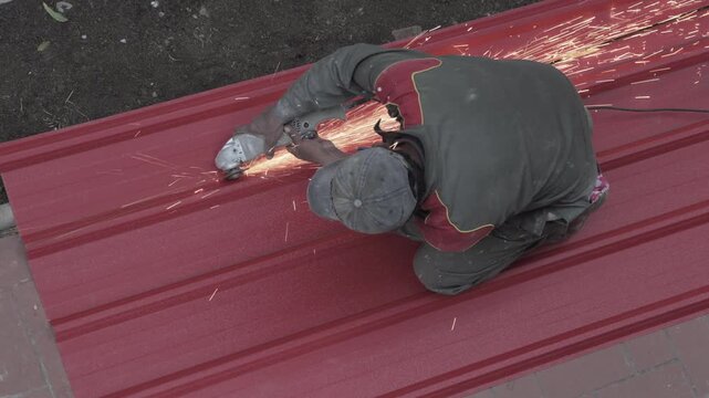 A bricklayer cutting a red corrugated metal sheet with a grinder, sending off golden sparks.