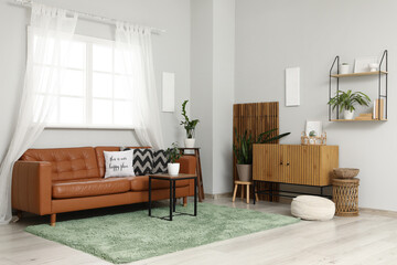 Fototapeta premium Interior of stylish living room with leather sofa, coffee table, houseplants and home decor on commode near window