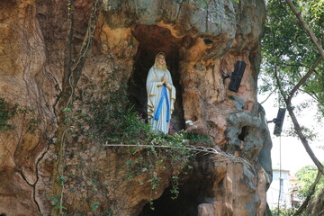 A serene statue of the Virgin Mary stands peacefully within a natural cave formation, surrounded by lush greenery and tree roots. Capturing a moment of spiritual reverence in nature
