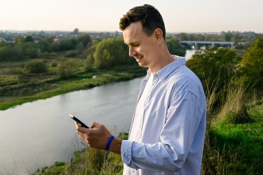 Young man in a shirt looking at smartphone with smile while standing by a riverside with scenic landscape in the background